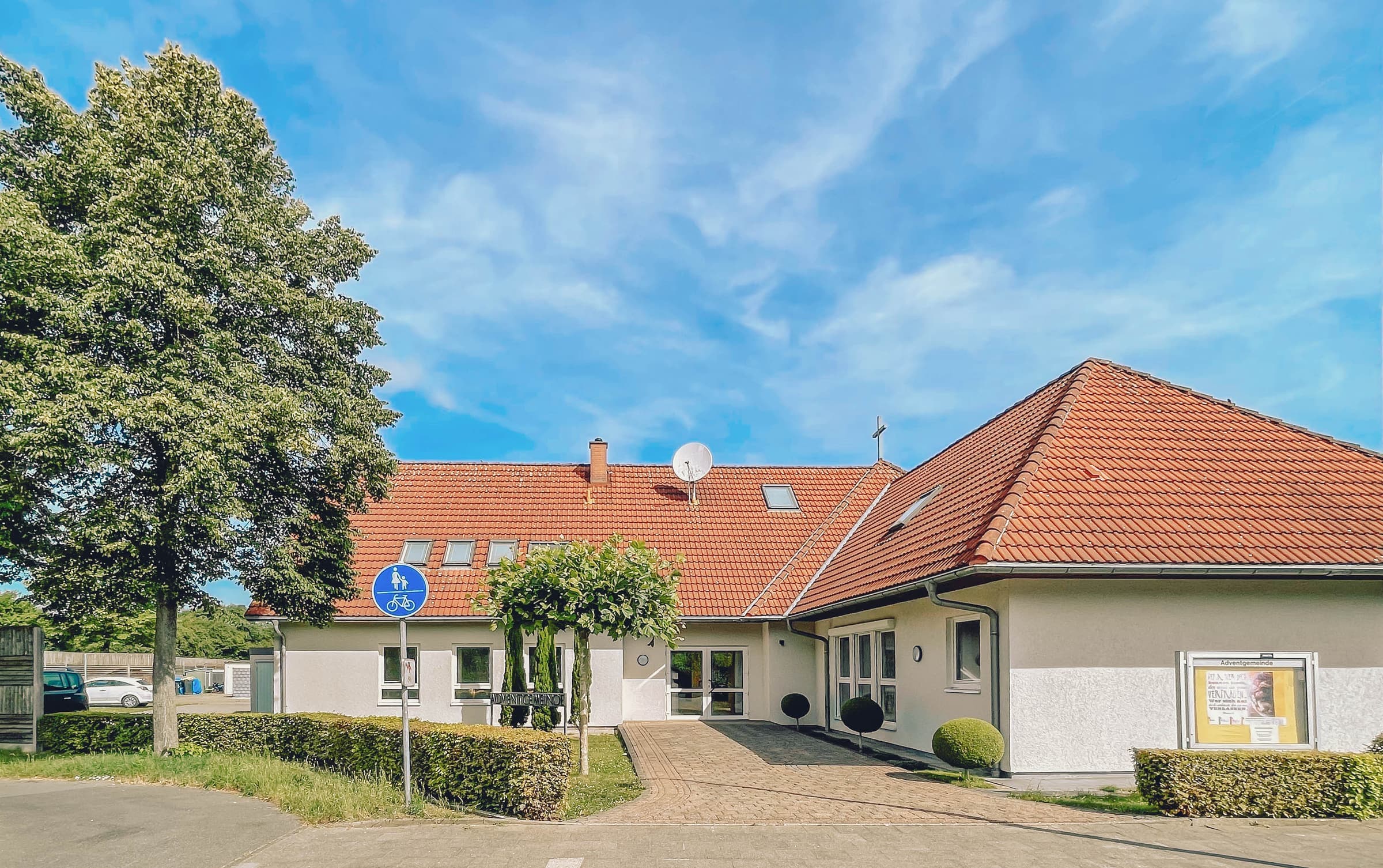 Das Gemeindehaus mit rotem Ziegeldach und gepflastertem Weg zum Eingang, davor ein großer Baum